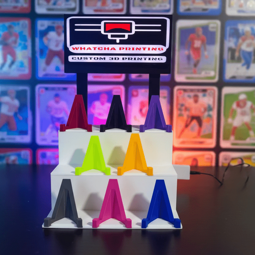 Colorful 3D-printed letter 'A's on a stand with 'Whatcha Printing' sign in the background.