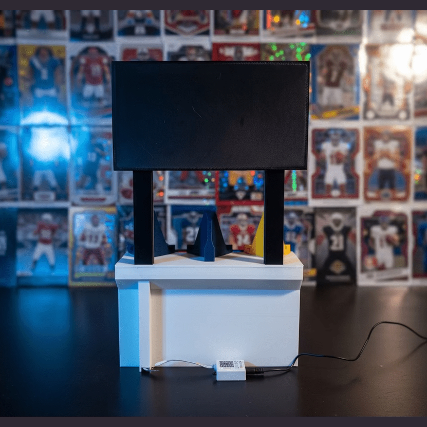 Small black screen on a white stand with a blue and orange object in front of it, on a dark surface.